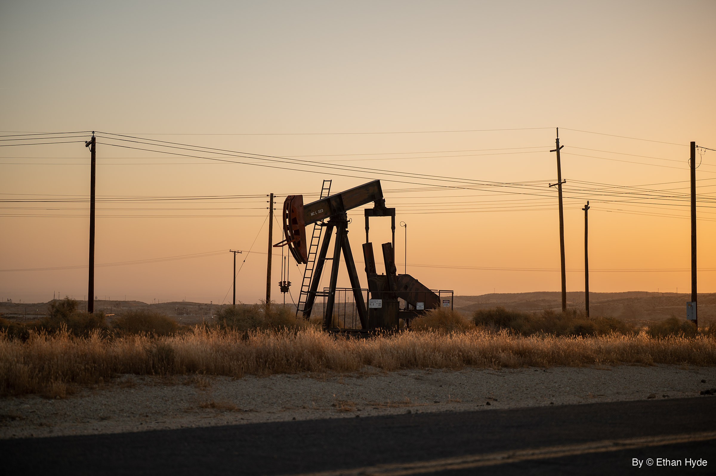 An oil pumpjack stands behind dry grass at the edge of a two-lane road, power lines stretching across a clear orange sunrise sky.