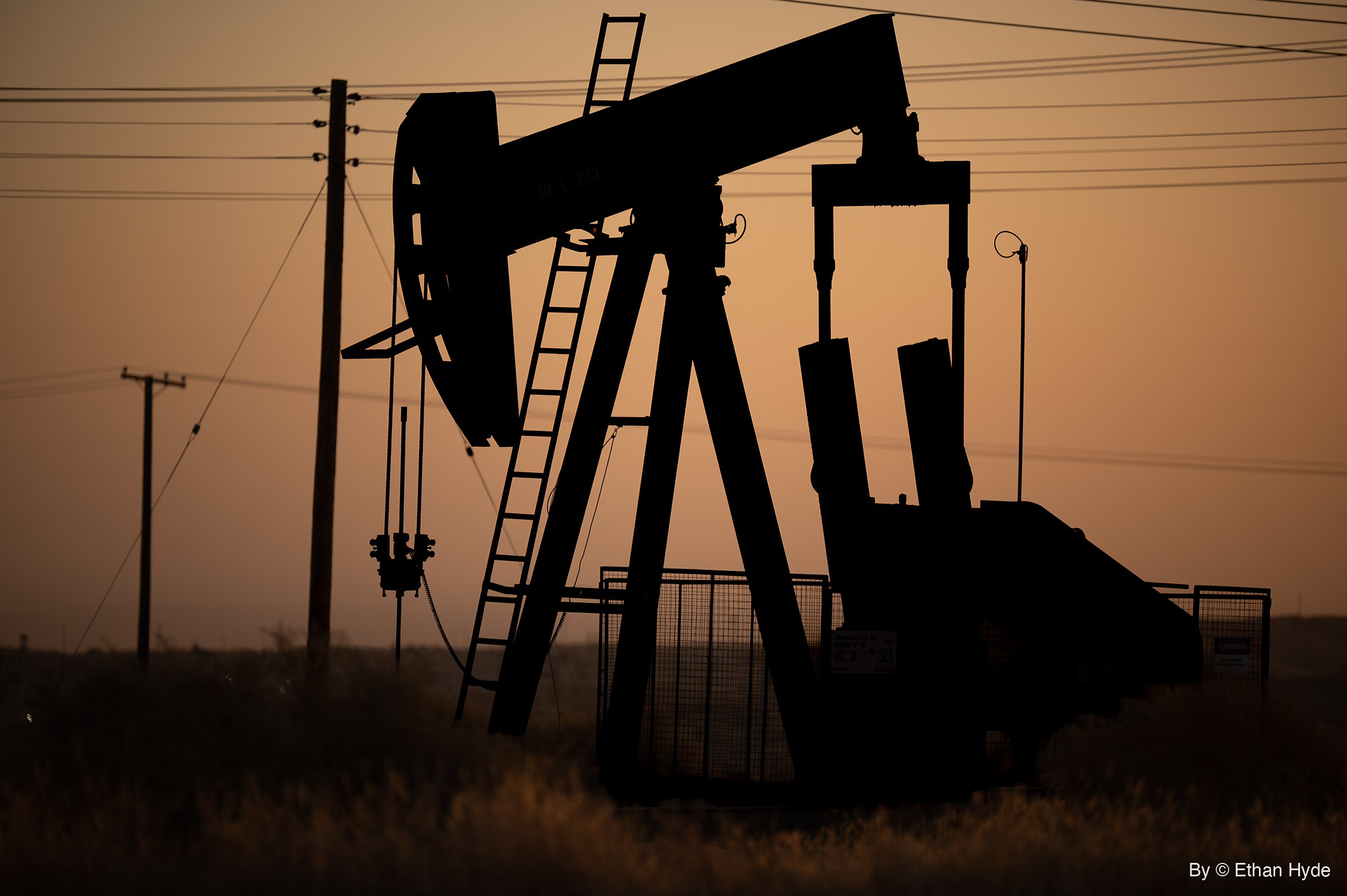 A close silhouette of an oil pumpjack with its ladder and counterweights against an orange sunrise sky, power lines and poles behind.
