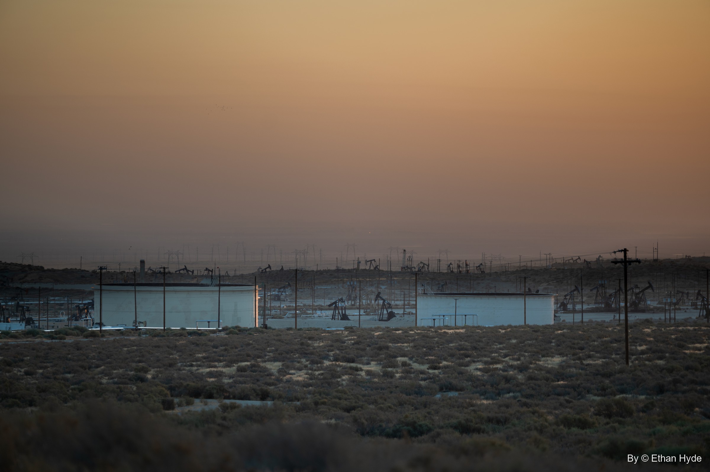 Rows of pumpjacks and storage tanks fade into amber haze across a flat at sunrise.