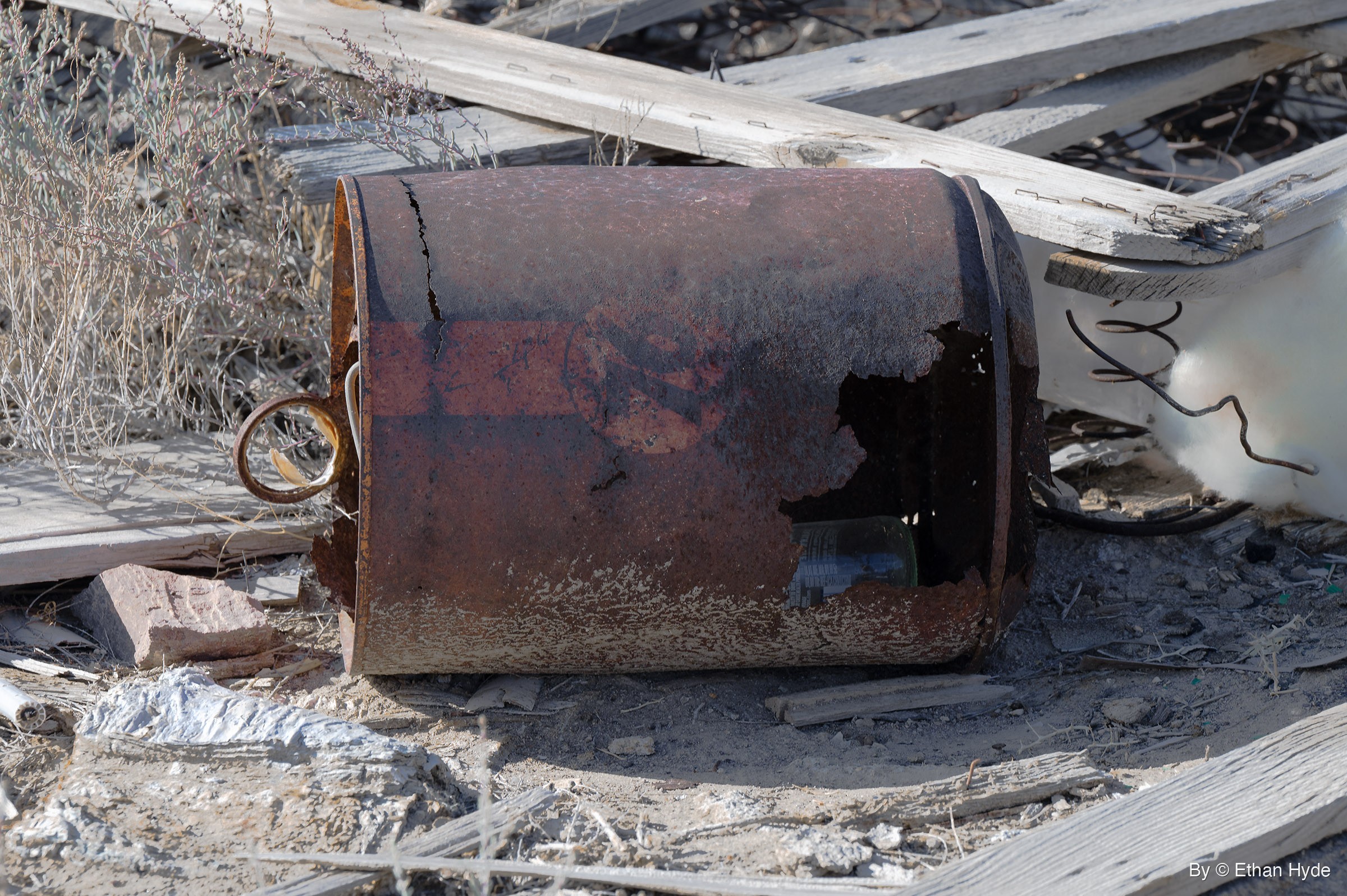 A rusted metal can with a ring handle lies on its side among weathered gray boards, coiled wire and dry brush, a glass bottle visible through a hole.