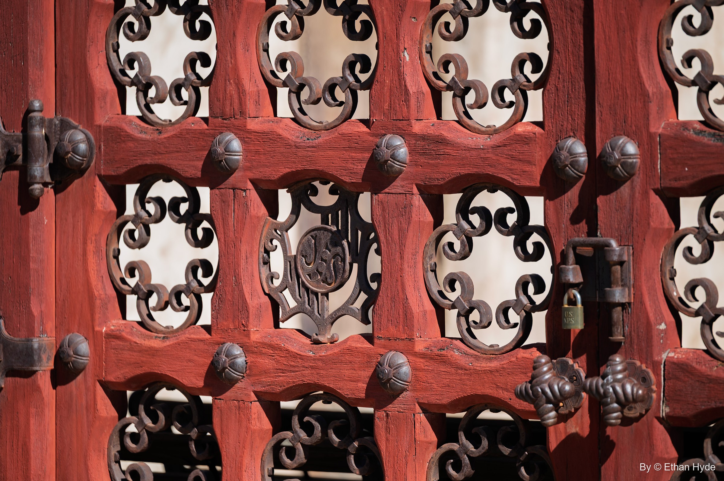 Close-up of the red courtyard gates at Scotty's Castle: iron scrollwork, studded rails, a padlock and a monogram panel.