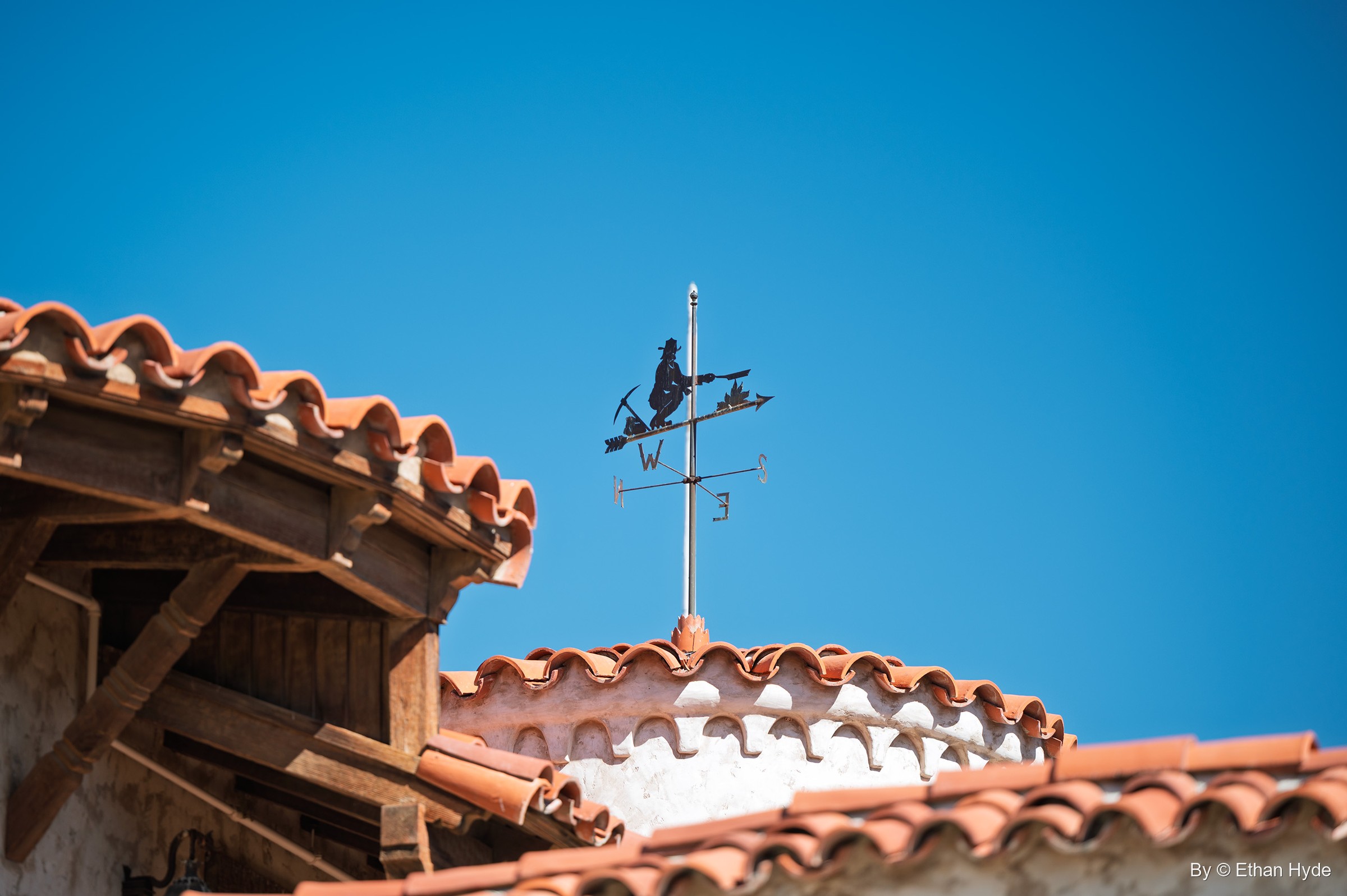 A weathervane in the form of a lone prospector figure above red tile roofs and timber eaves at Scotty's Castle.