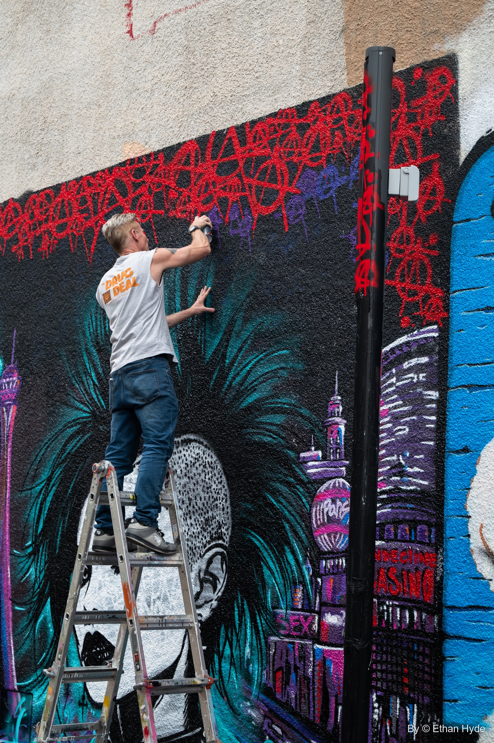 An artist on a ladder paints a large portrait with a city skyline and observation tower, beneath a band of red scribbles.