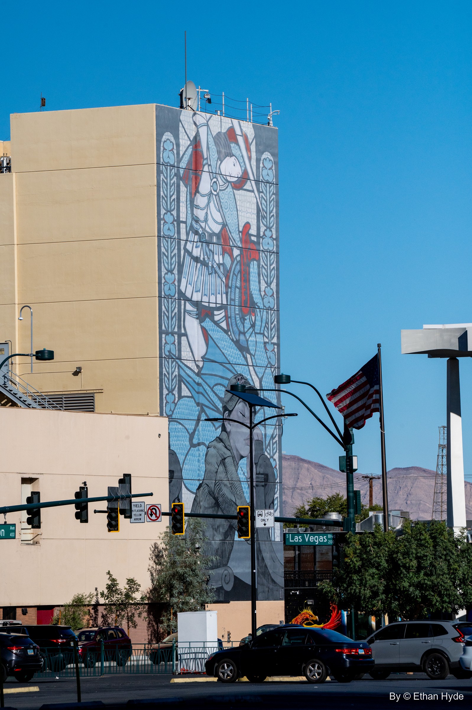 A mural in a stained-glass style covers a tall building wall above traffic lights, an American flag and desert mountains.