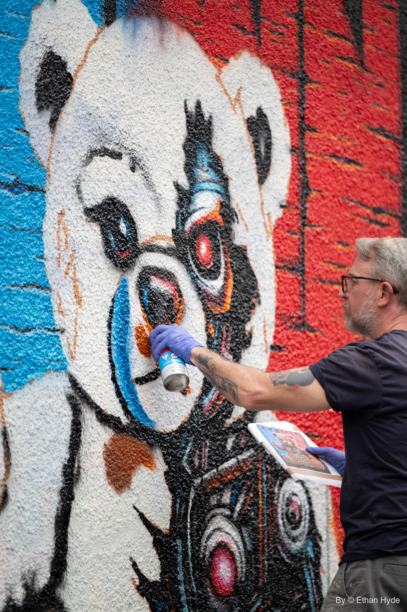 A gray-haired artist in blue gloves sprays a mural of a teddy bear holding a camera against a red brick-pattern background.