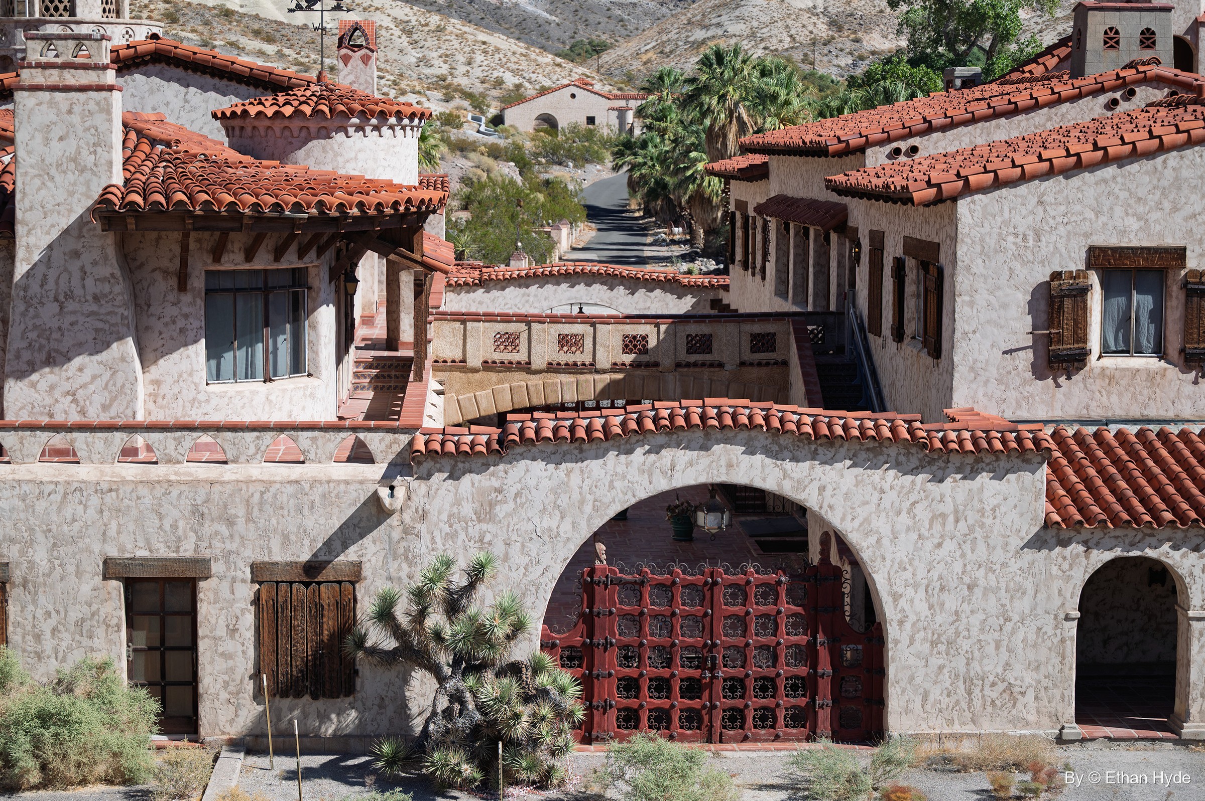 The main facade of Scotty's Castle: an arch with red wood-and-iron gates flanked by stucco wings, a Joshua tree and palms beyond.