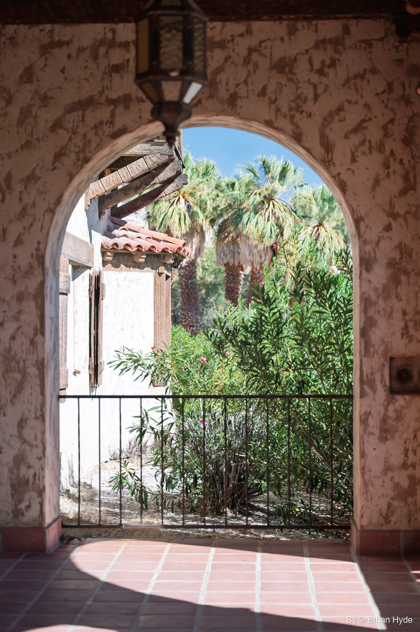 A view through a stucco courtyard arch at Scotty's Castle to palms and a tiled roof, a lantern hanging overhead and red tile underfoot.