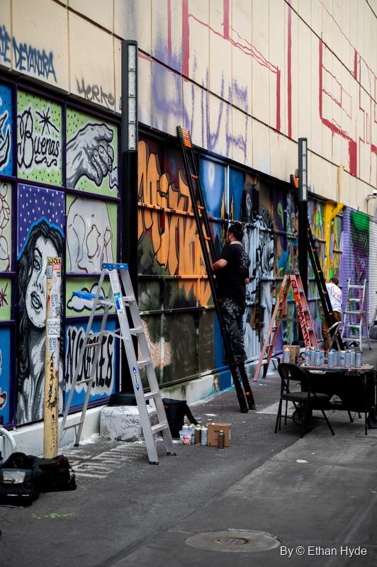 A long wall of murals in progress, with artists up ladders and a folding table of spray cans along the sidewalk.