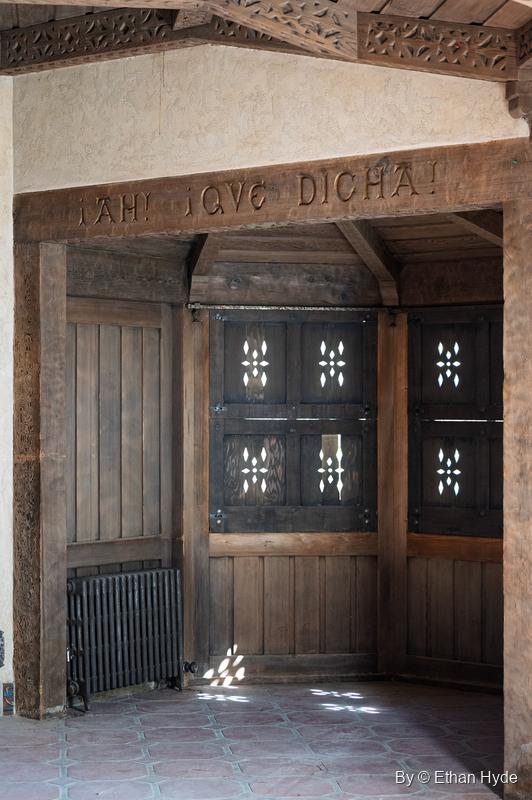 A carved wooden beam reading "¡Ah! ¡Que dicha!" over a wood-panelled alcove with pierced wooden doors at Scotty's Castle.