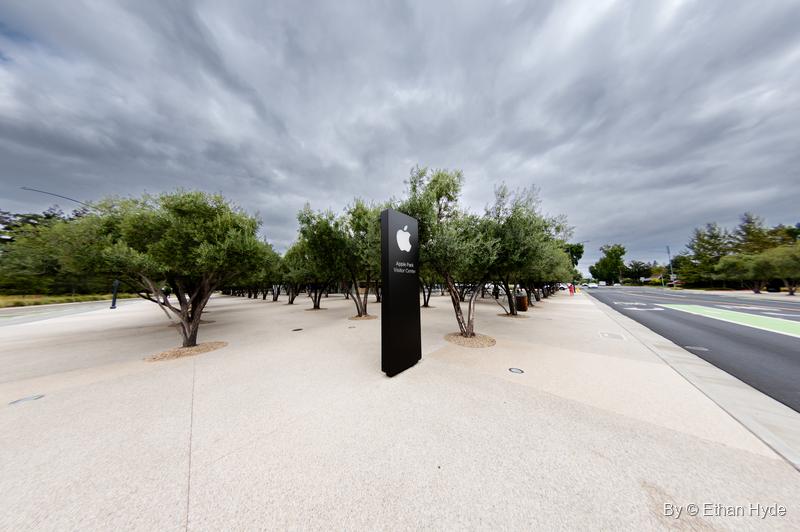 A black Apple logo sign stands among rows of olive trees on a pale plaza beside the road, under gray clouds.