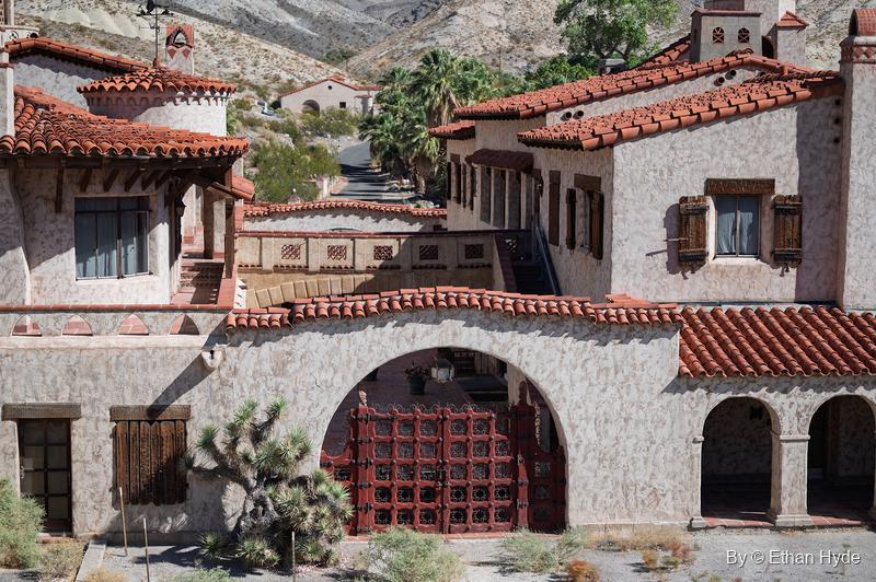 Stucco facade of Scotty's Castle with a red-gated archway and a Joshua tree, rooftops stepping back toward the canyon.
