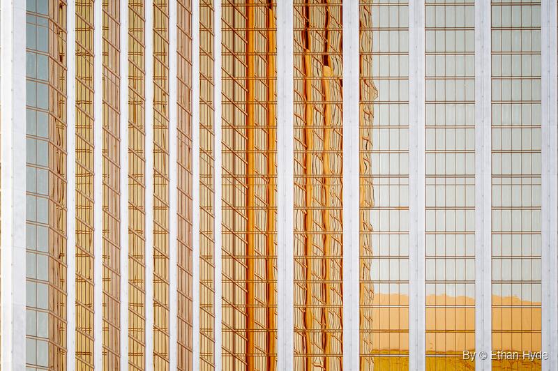 A tall building facade with white vertical columns and gold-tinted windows reflecting warped shapes of neighboring towers.