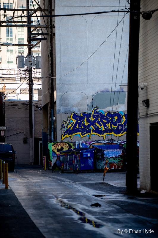 A narrow alley framed by power poles opens onto a blue and yellow graffiti piece over tagged dumpsters.