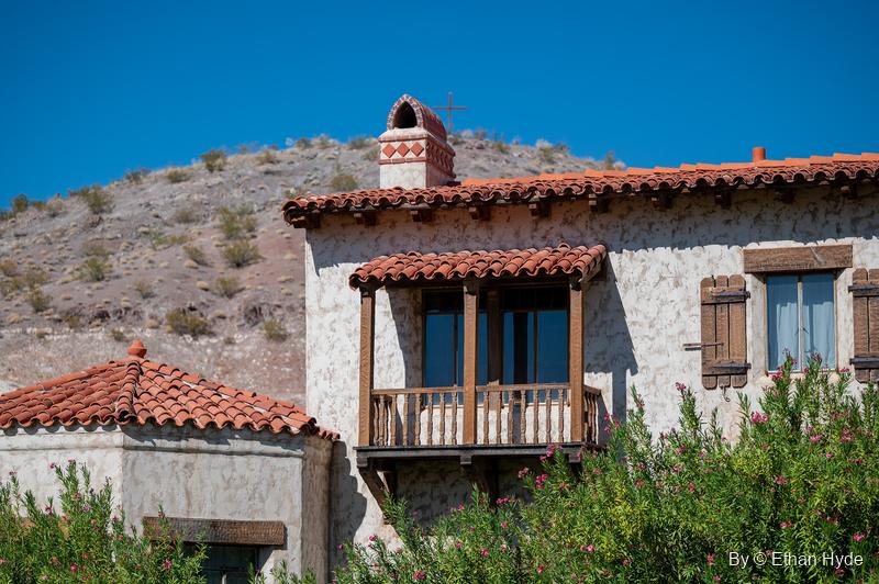 A wooden balcony under a red tile roof on a stucco wing of Scotty's Castle, with flowering oleanders and a desert hillside behind.