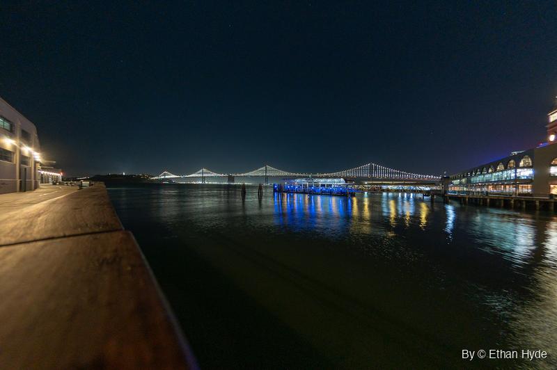 The lit San Francisco-Oakland Bay Bridge stretches across dark water at night, with a lit pier building on the right and a wooden railing leading in from the left.