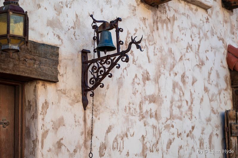 A small bell hangs from a scrolled wrought-iron bracket on a stucco wall at Scotty's Castle, a lantern at the edge of the frame.