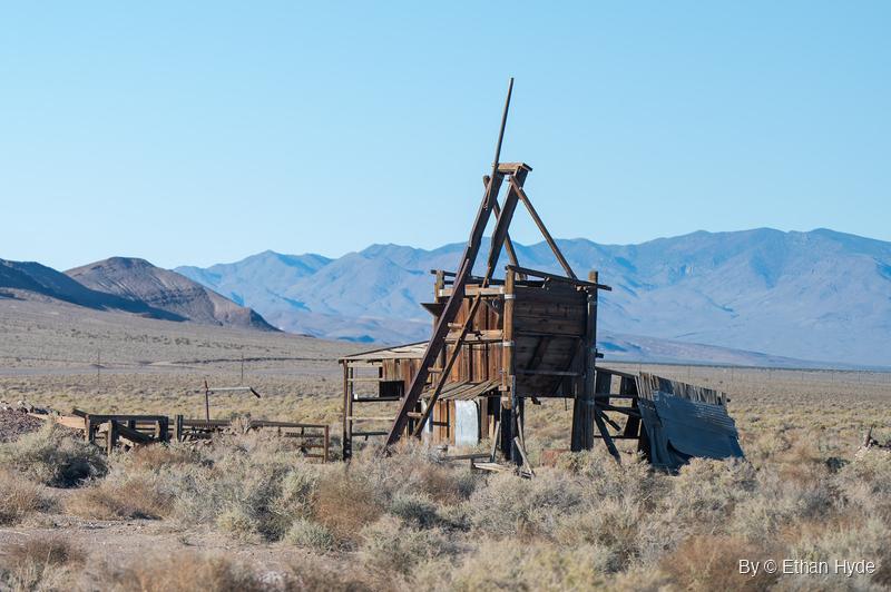 A weathered wooden headframe with a tall leaning pole stands in sagebrush, with blue mountain ranges and a line of poles behind it.