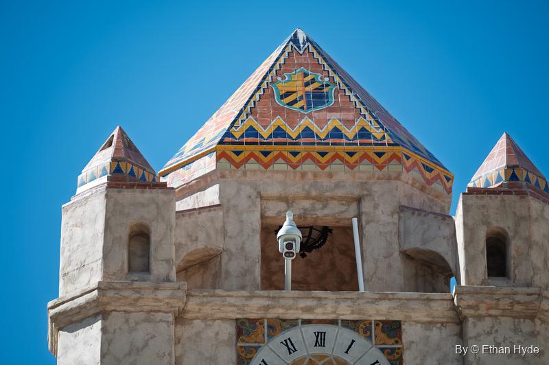 The tiled pyramid roof of the Chimes Tower at Scotty's Castle, with a coat of arms, the open chime chamber and the top of the clock face.
