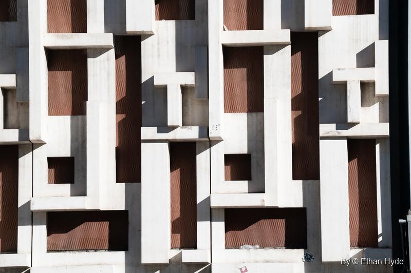 Abstract close-up of a white concrete facade of interlocking raised blocks over dark brown recesses.