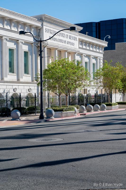 The full white facade of the Nevada Supreme Court building behind young trees and a black street lamp, seen across an empty street.
