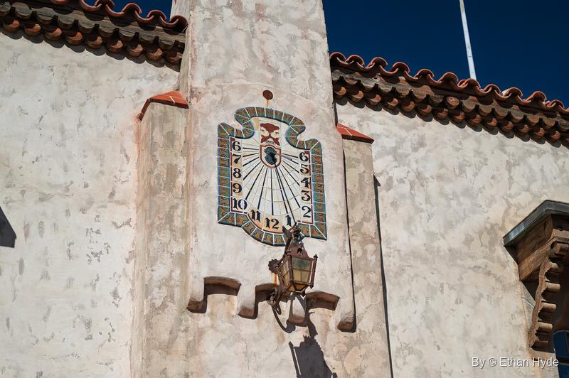 A painted sundial with numbered hours mounted high on a stucco wall of the main house at Scotty's Castle, with a lantern below it.