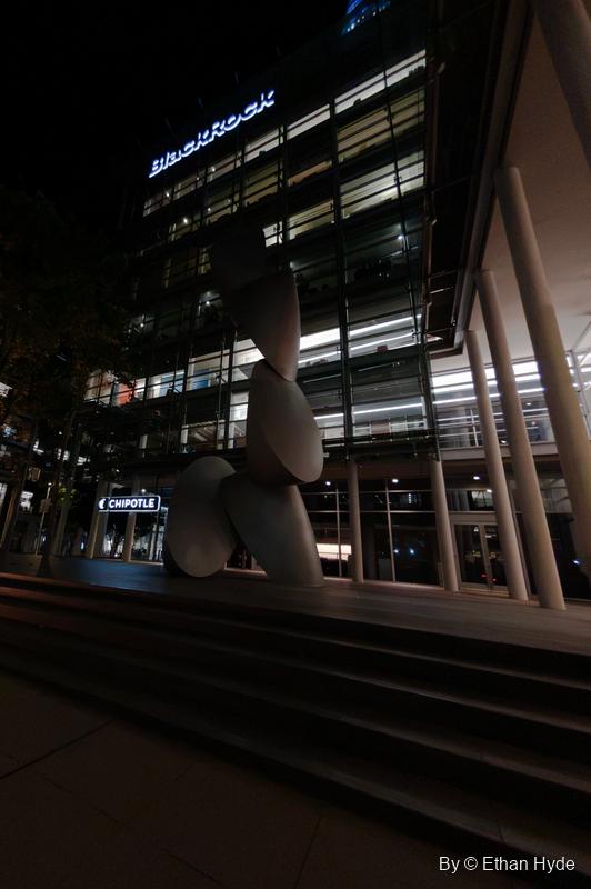 Close vertical view of the stacked disc sculpture Time Signature in front of a lit office facade at night.