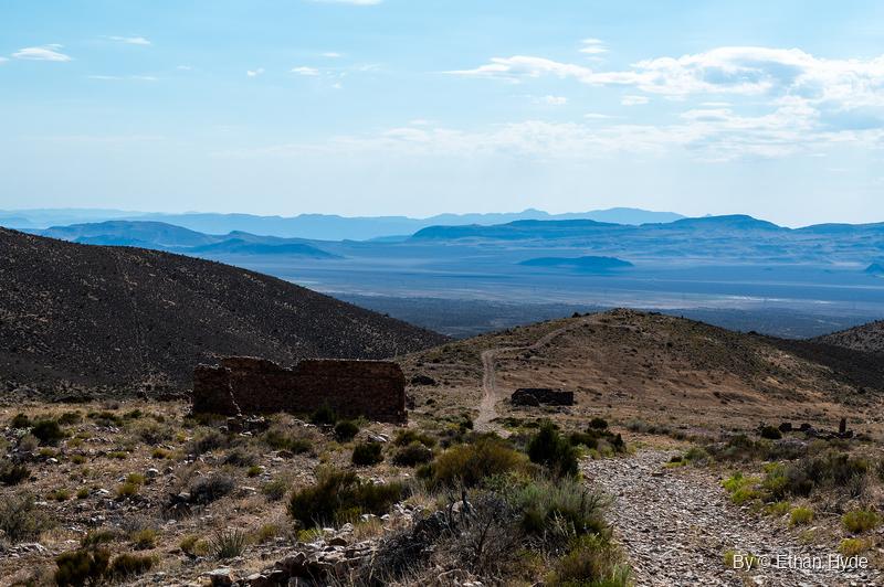 Roofless stone walls of Delamar ruins sit on a scrubby hillside beside a rocky track, with a wide valley and hazy blue ranges beyond.