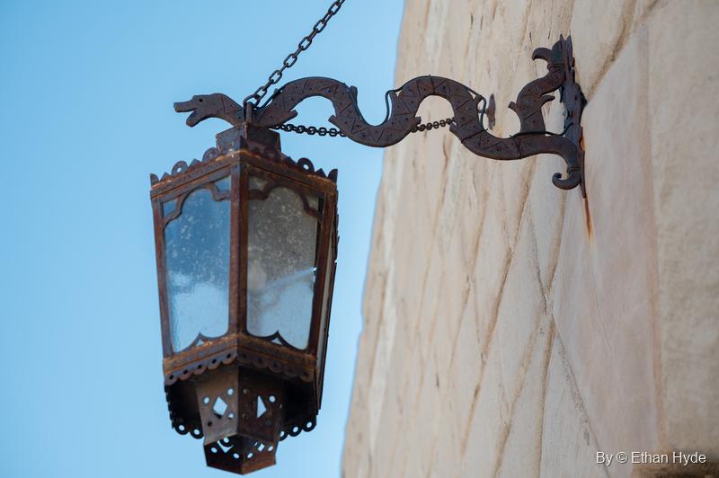 A wrought-iron lantern hangs from a scrolled beast-head bracket on a stone wall at Scotty's Castle, against a pale blue sky.