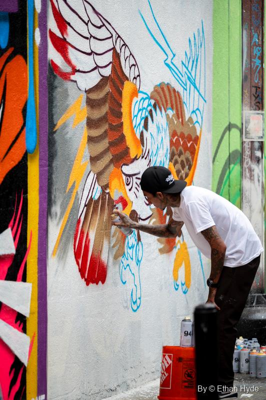 An artist in a cap sprays a mural of an eagle with lightning bolts, paint cans in an orange bucket at his feet.