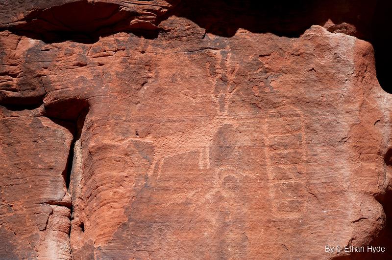 Weathered red sandstone wall with a faint pecked animal figure and a grid-like shape beside it, below a shadowed overhang.
