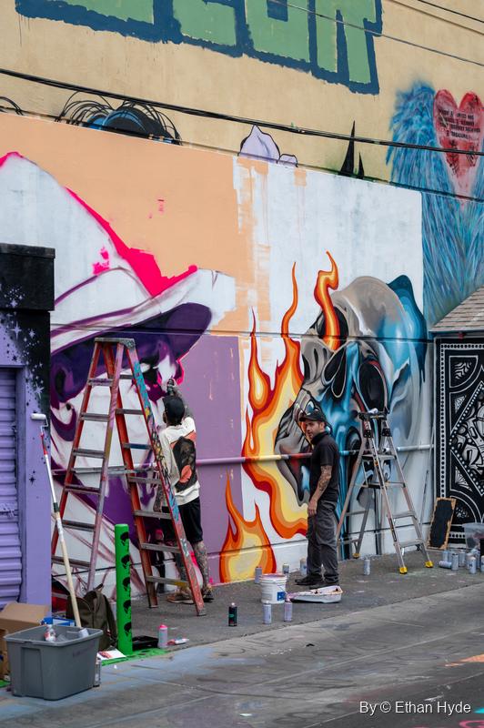 Two painters on ladders work on a mural of flames and a blue skull against a purple and peach wall.