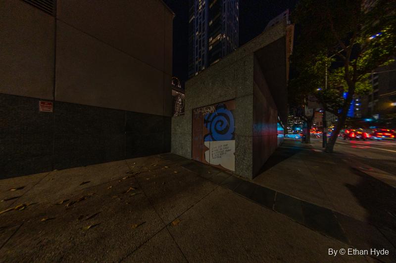 At night, a boarded doorway in a granite wall is painted with a blue spiral and the words "Let's fit our wings in this collective home & fly free together."