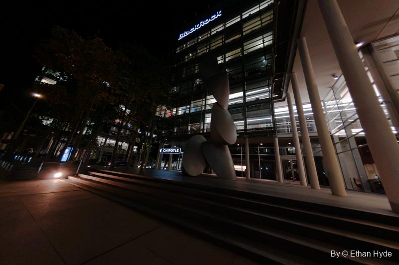 Wide night view of broad steps, angled columns and a stacked white sculpture before a glass office building.