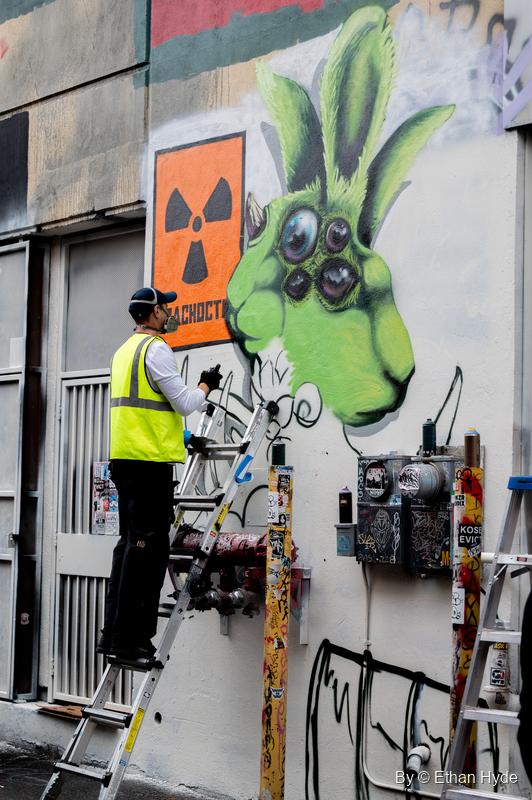 A painter in a hi-vis vest stands on a ladder spraying a green four-eyed rabbit creature beside an orange radiation-symbol panel.
