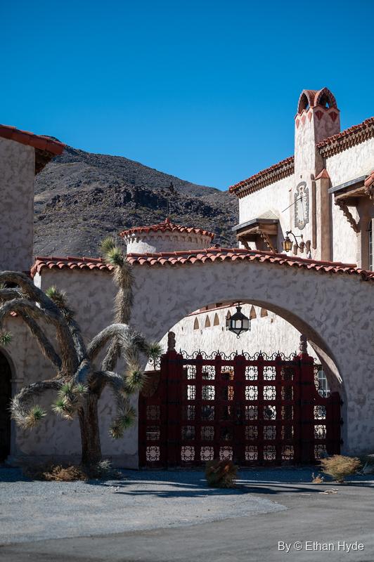 A Joshua tree frames the red-gated archway of Scotty's Castle, a hanging lantern inside the arch and dark hills above.