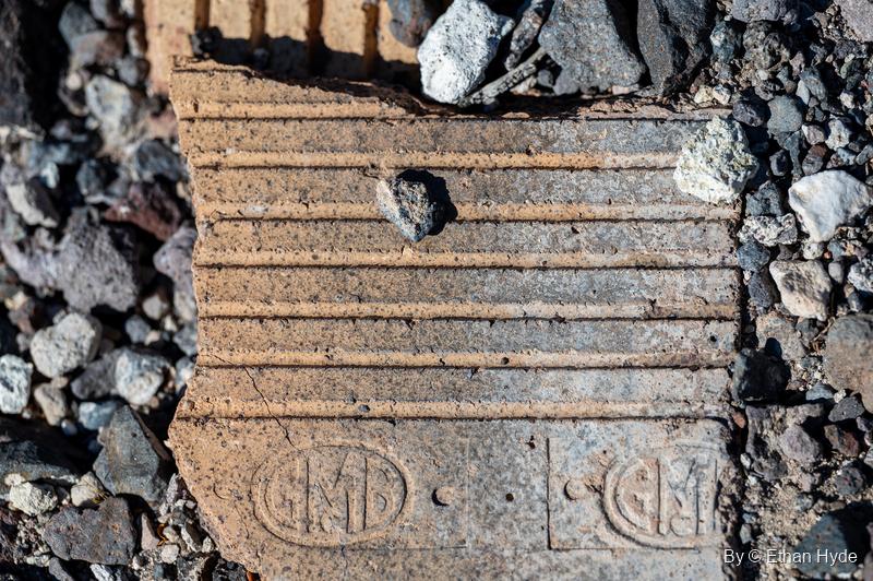 A broken ribbed clay block stamped with an oval "GMB" monogram, lying in volcanic gravel near Scotty's Castle.