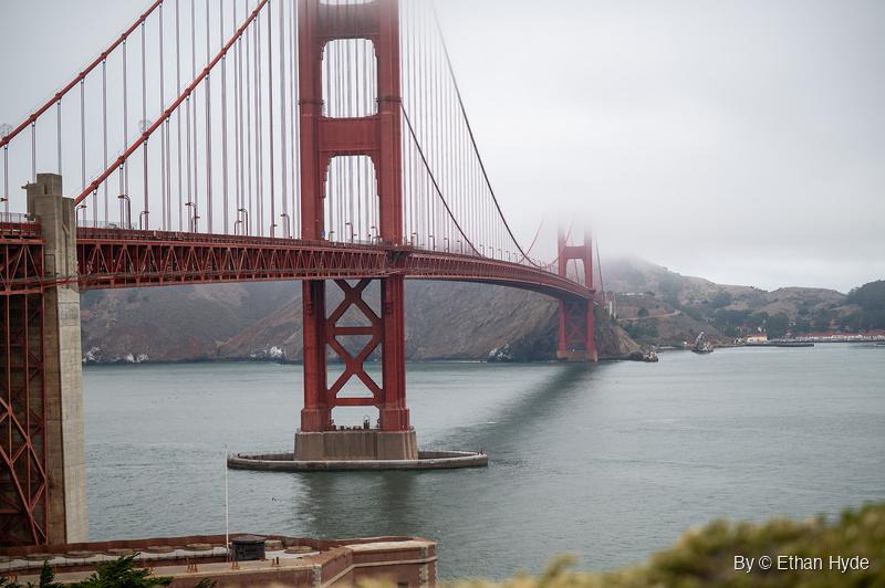 The Golden Gate Bridge's red towers rise over gray water, the far tower's top lost in low fog above the Marin Headlands.