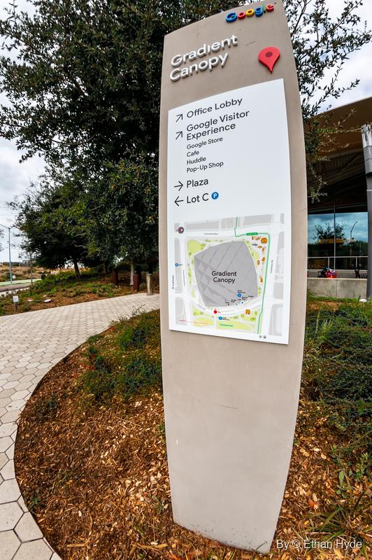 A curved wayfinding sign for Google's Gradient Canopy with a campus map, beside a path and mulched planting.