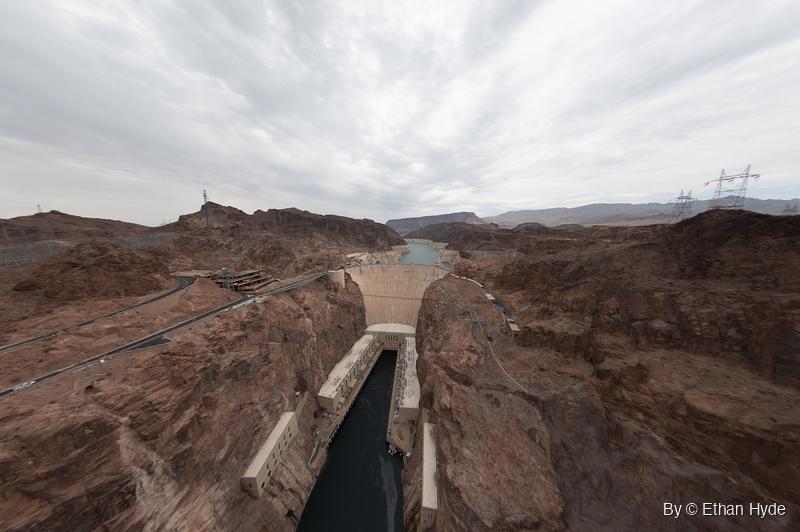 Ultra-wide view of Hoover Dam in its rock canyon, with curving highways on the left and transmission towers on the ridges under an overcast sky.