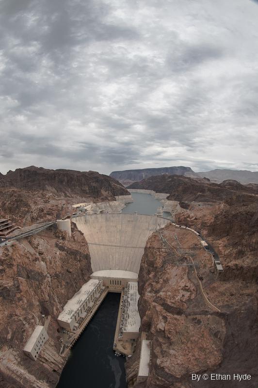 Vertical fisheye view down onto Hoover Dam, its powerhouses and the dark river below, with the reservoir and a mesa behind under heavy cloud.