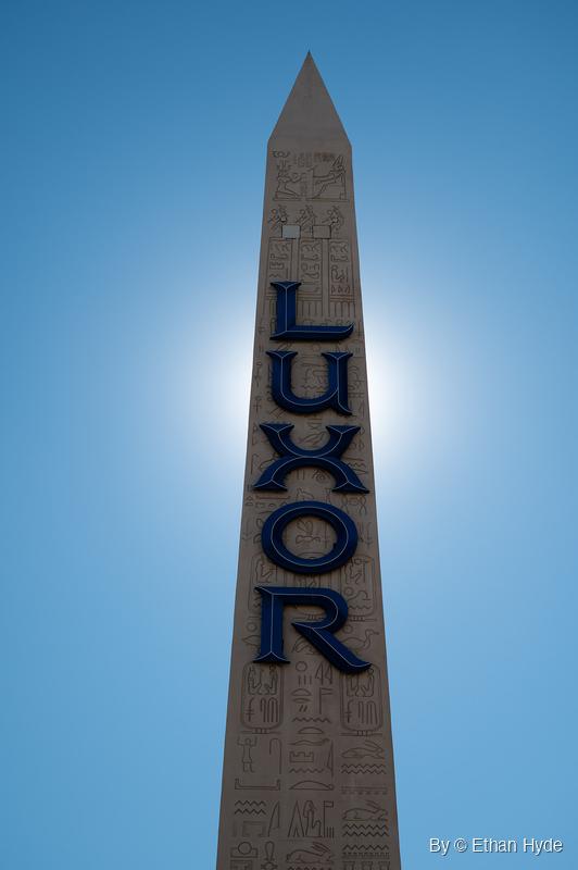 Tall Egyptian-revival obelisk with carved hieroglyph panels and large vertical lettering, seen from below with the sun directly behind it.