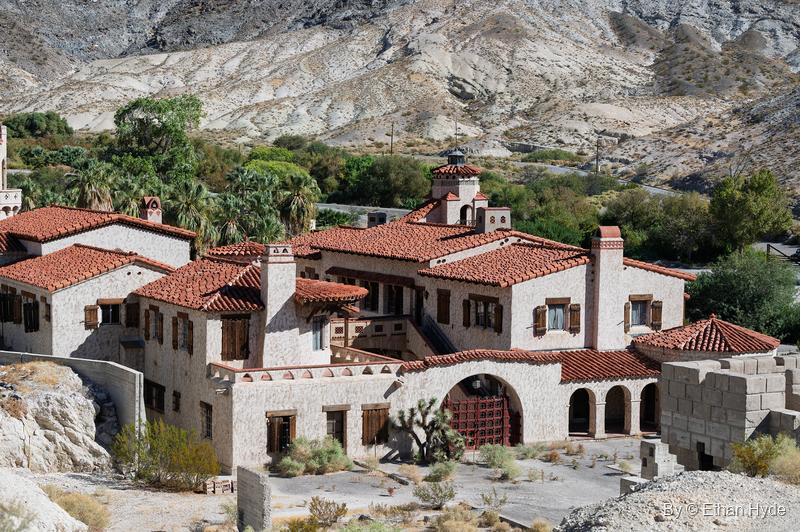 An elevated view of the main house and annex of Scotty's Castle, red tile roofs and stucco walls set against the oasis trees and pale hills.