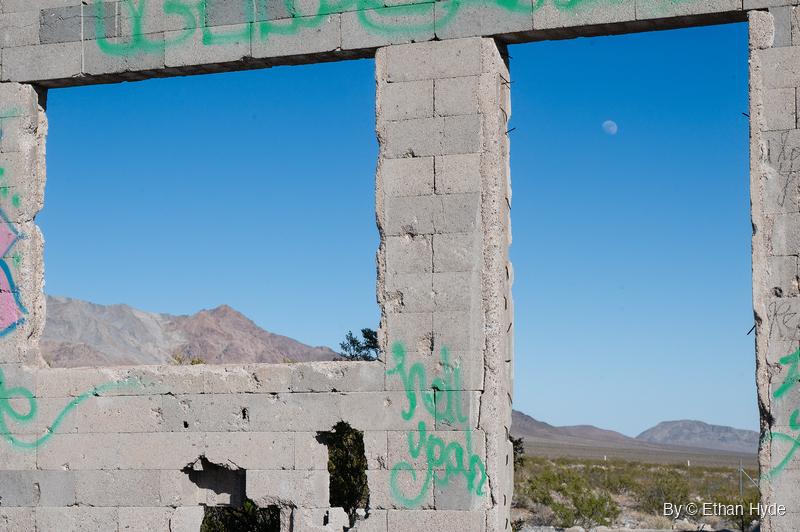 A roofless graffiti-covered cinderblock building frames blue sky, a daytime moon and desert mountains through its empty window openings.