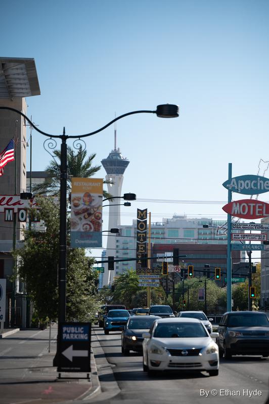 A downtown street of motel signs, palms and street lamps, with a tall observation tower rising in the distance.