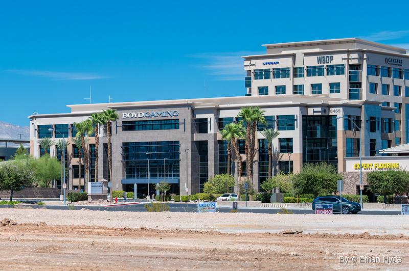 A four-story granite and glass office building with a taller office tower behind it, palm trees along the street and a dirt lot in front.