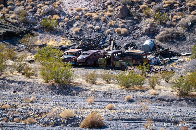 Rusted vintage cars and a truck parked among creosote bushes and scrap lumber on a rocky slope near Scotty's Castle.