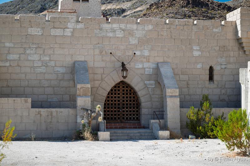 A crenellated stone-block wall at Scotty's Castle with a pointed-arch iron grille doorway and a hanging lantern.