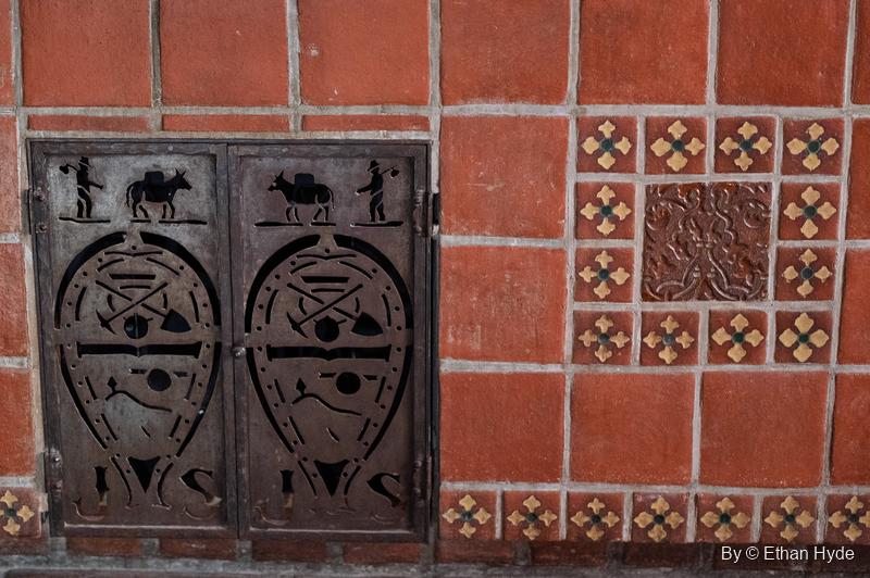 A pair of small iron doors in red kitchen tile at Scotty's Castle, cut out with a prospector and burro, a pick and shovel, and the initials J and S.