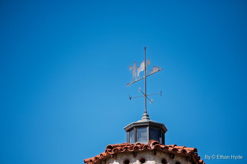 A weathervane showing a prospector carrying a bundle and leading a pack burro, atop a cupola at Scotty's Castle against a blue sky.