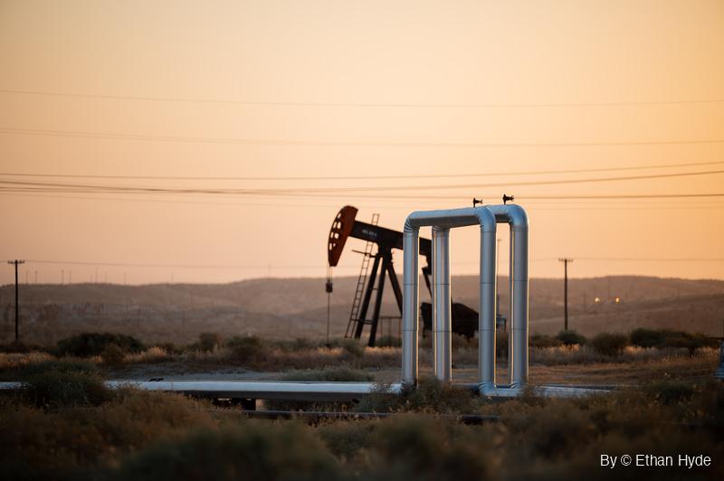 An oil pumpjack and a raised silver pipe loop stand in dry brush under an orange sunrise sky crossed by power lines, with low hills behind.